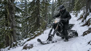 Helmeted rider guiding a tracked snow motorcycle along a narrow, snow-covered forest trail beside a steep wooded slope.